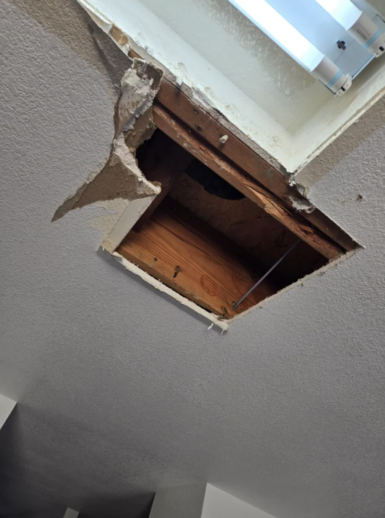 New York City Ceiling with a square hole exposing wooden framing and a vent pipe, with some drywall edges broken. Before ceiling repair