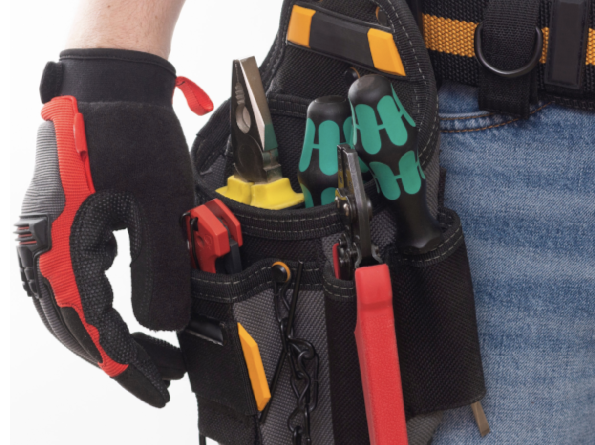 Person wearing work gloves with a tool belt holding various tools including pliers, screwdriver, and a wrench.