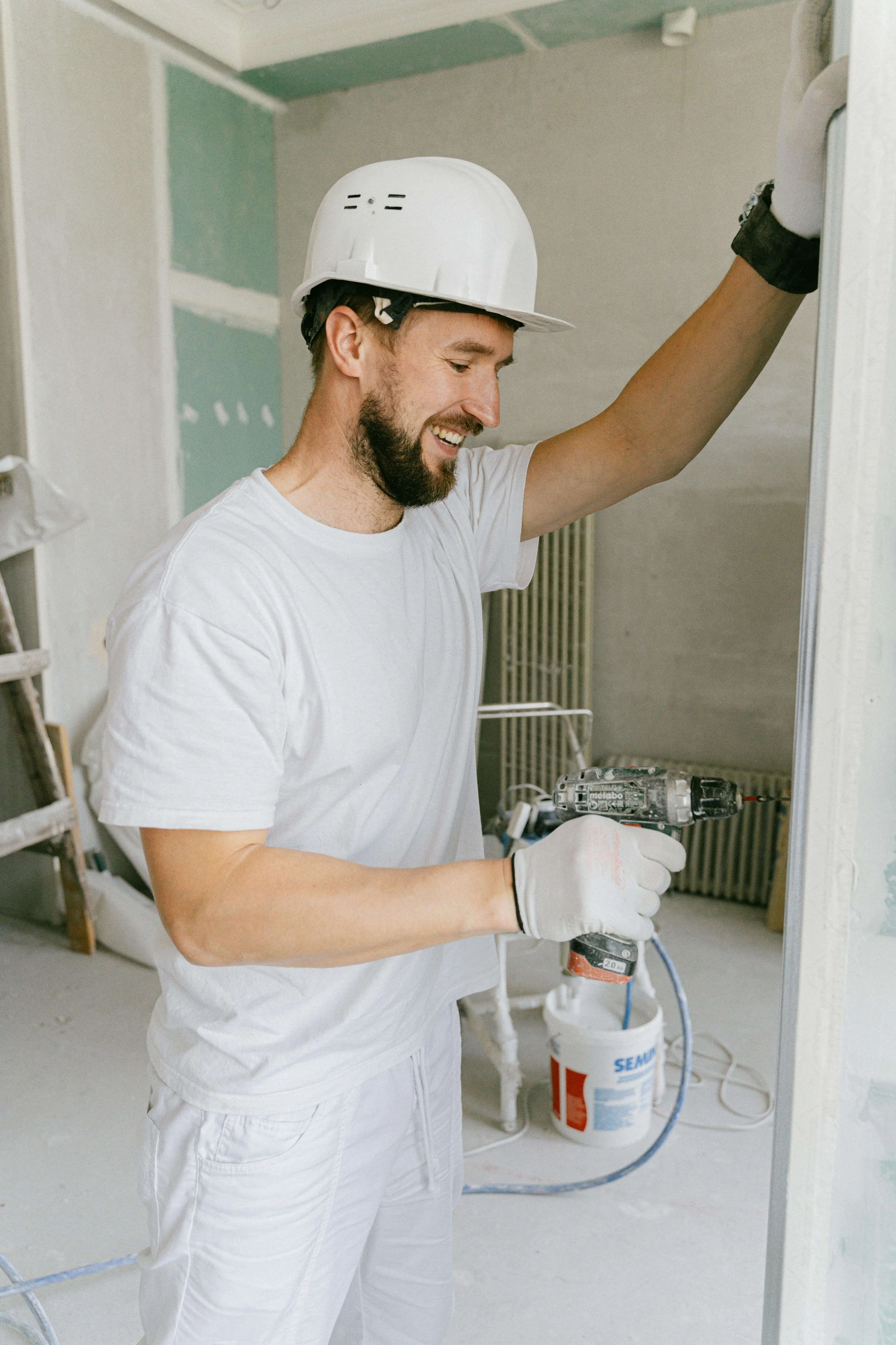A man wearing a white hard hat and white clothing is smiling while using a cordless drill to install a frame on a wall in a room under construction.