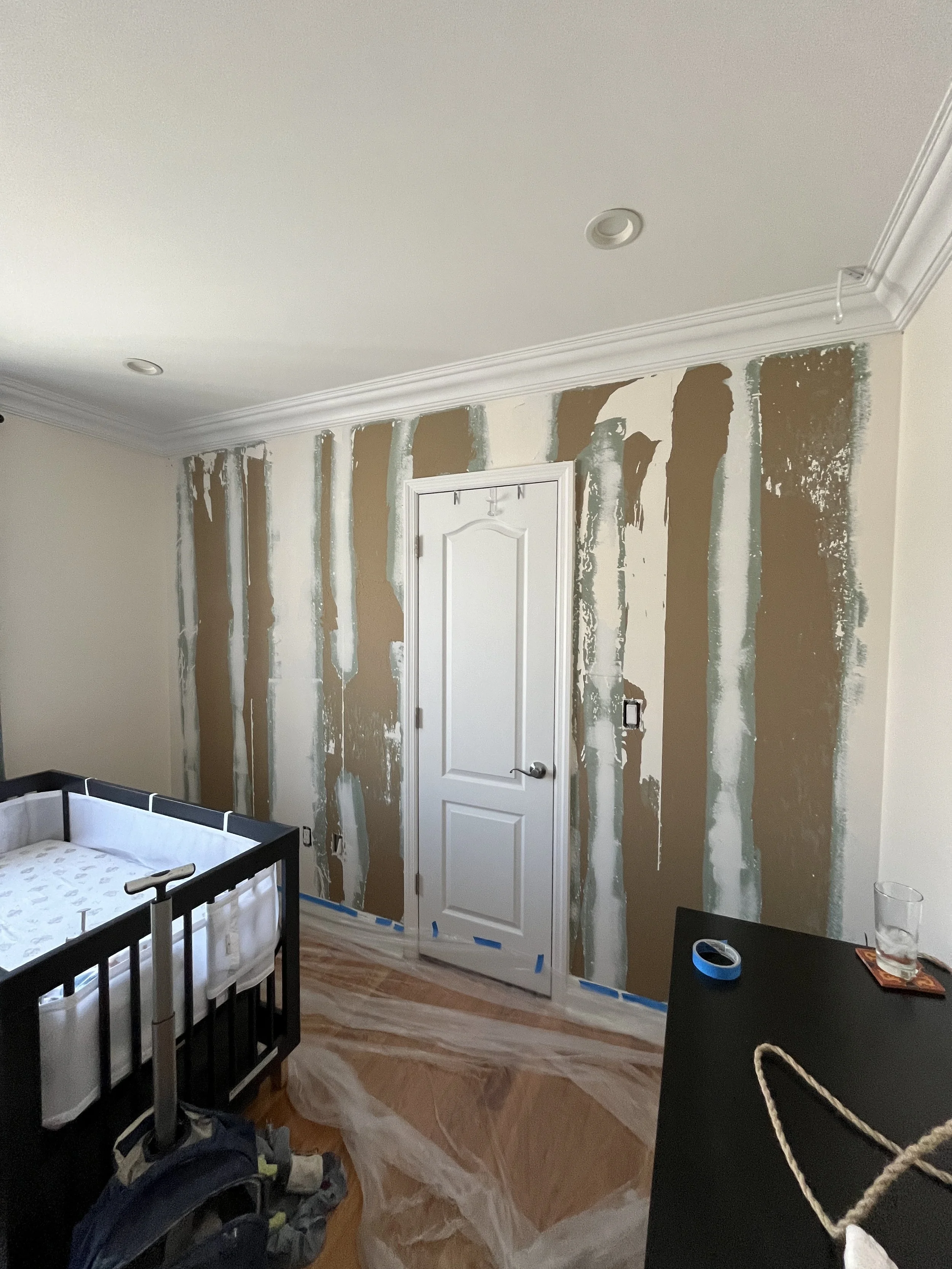Room with a door currently being painted, with visible tape and patchy paint on the walls, a crib, and a black dresser with miscellaneous items.