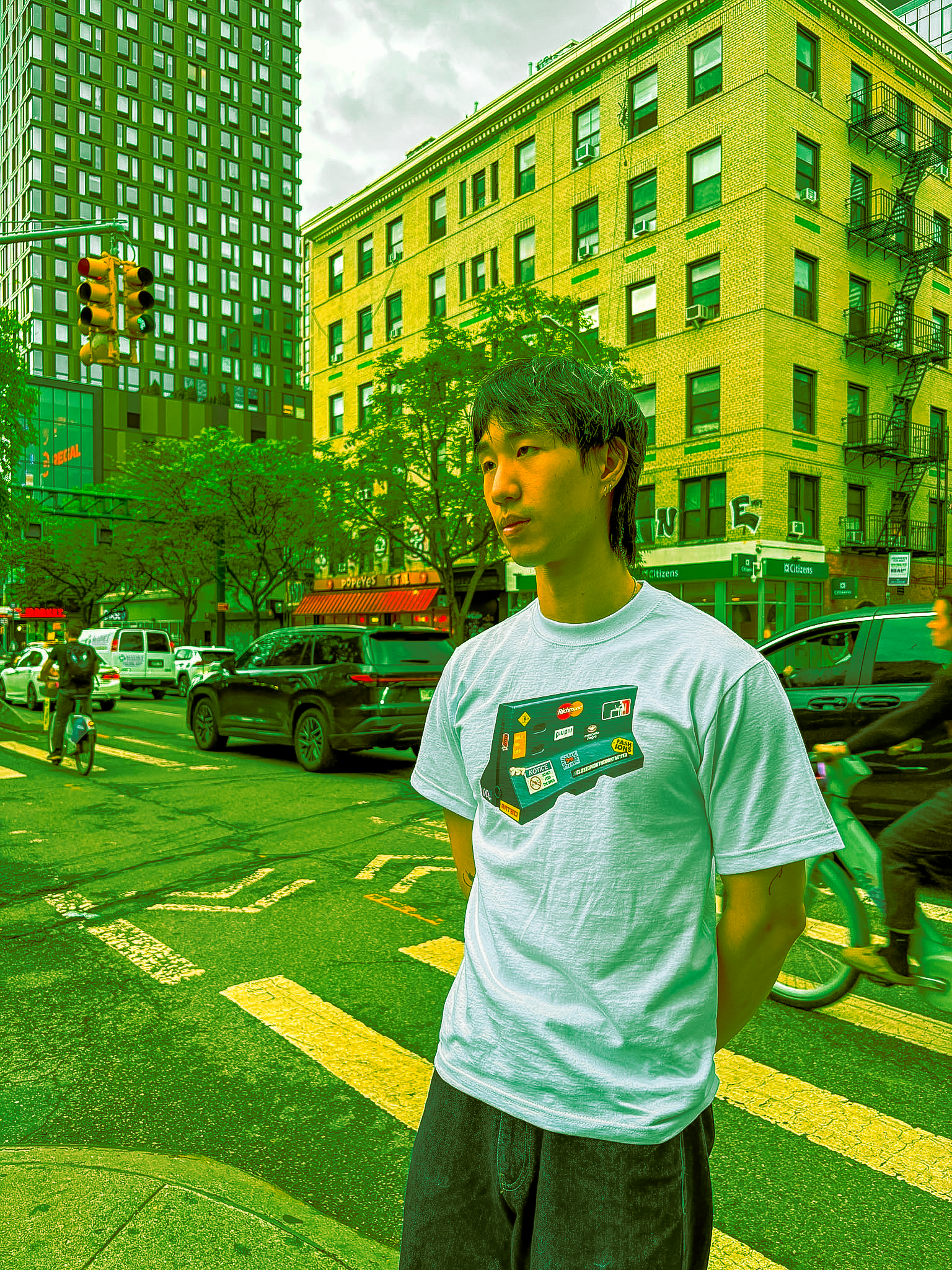 A young person with dark, spiky hairstyle standing on a city street with tall buildings, traffic lights, and cars in the background. They are wearing a white t-shirt with various logos and black pants, and are standing with their hands behind their b