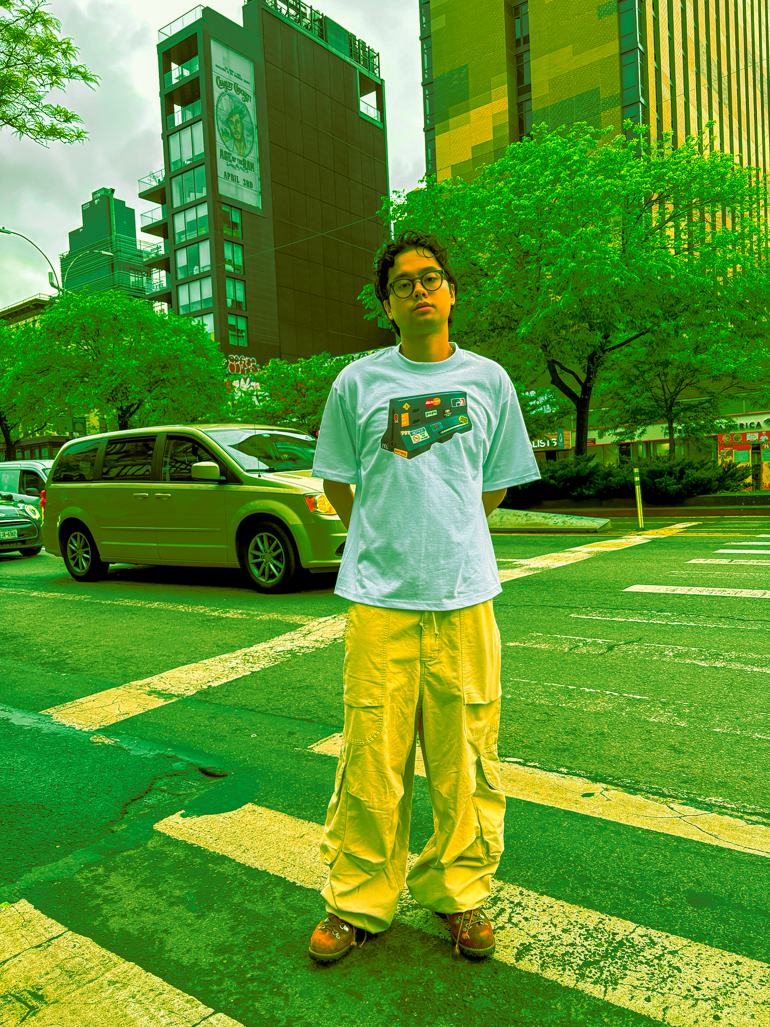 A young man with curly hair, glasses, and a serious expression standing in a city street with cars, green trees, and modern buildings in the background. He is wearing a white T-shirt with stickers and yellow cargo pants.