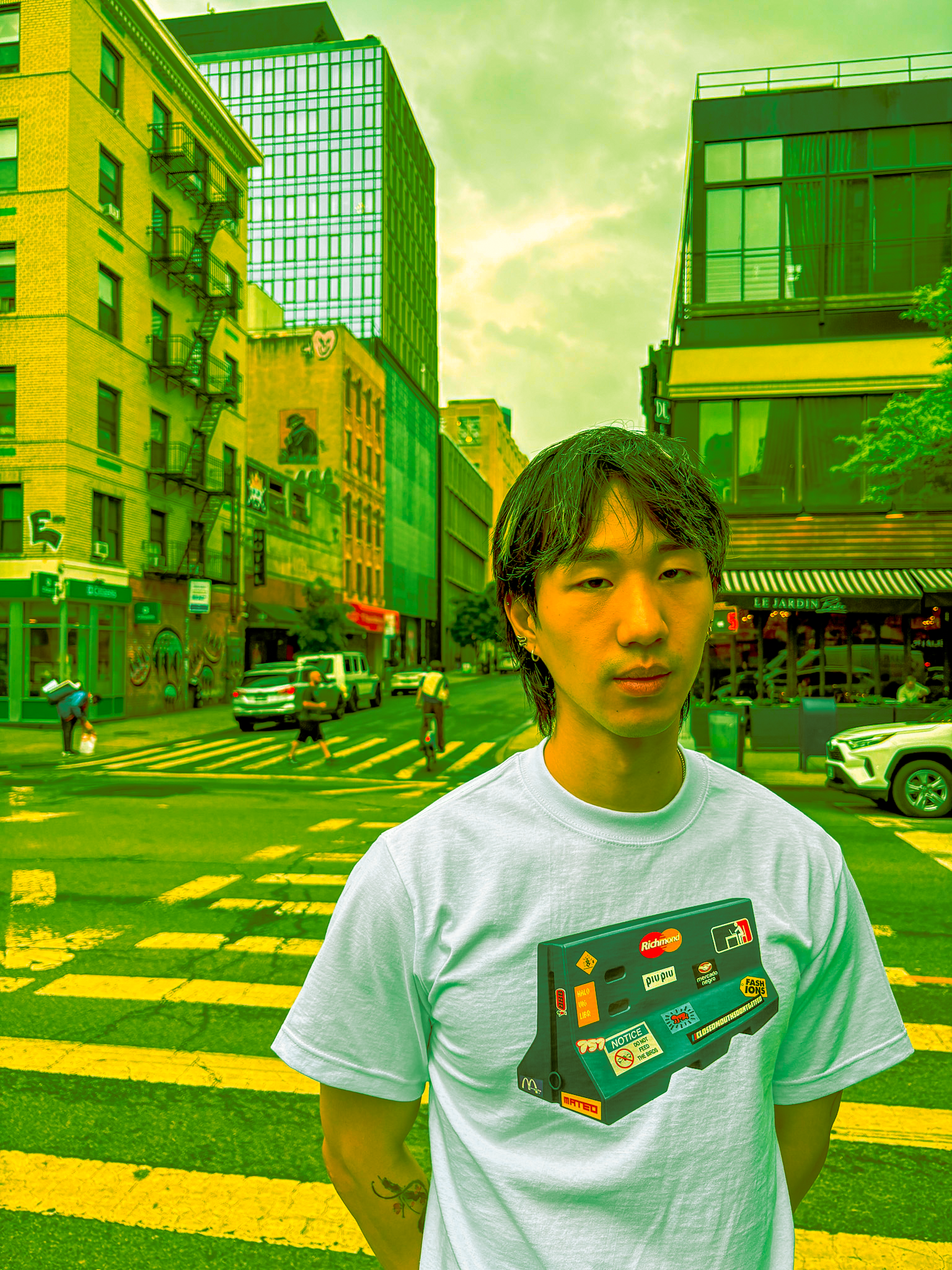 A young man with dark, wet hair wearing a white T-shirt stands on a city street at sunset. He is holding a black rectangular object with various stickers on it, including logos and warning signs. The background features tall buildings, some with glas