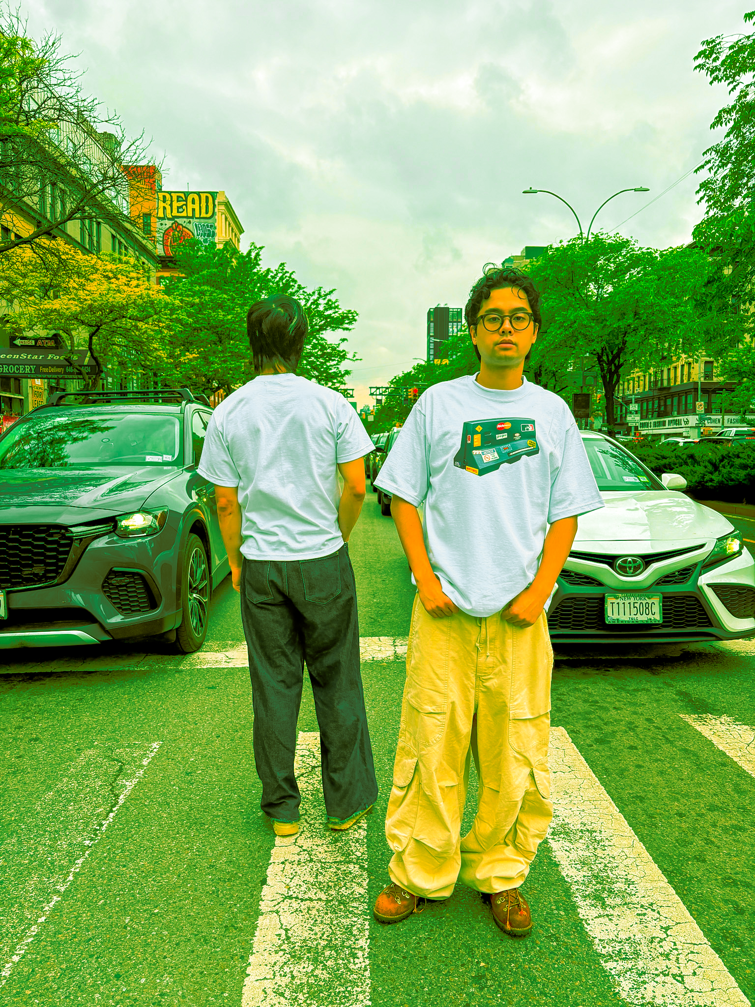 Two young men standing on a crosswalk on a city street, with cars parked on either side. One faces forward with glasses and casual clothes, the other turns away. Trees and buildings line the street, with a billboard reading "READ" in the background.