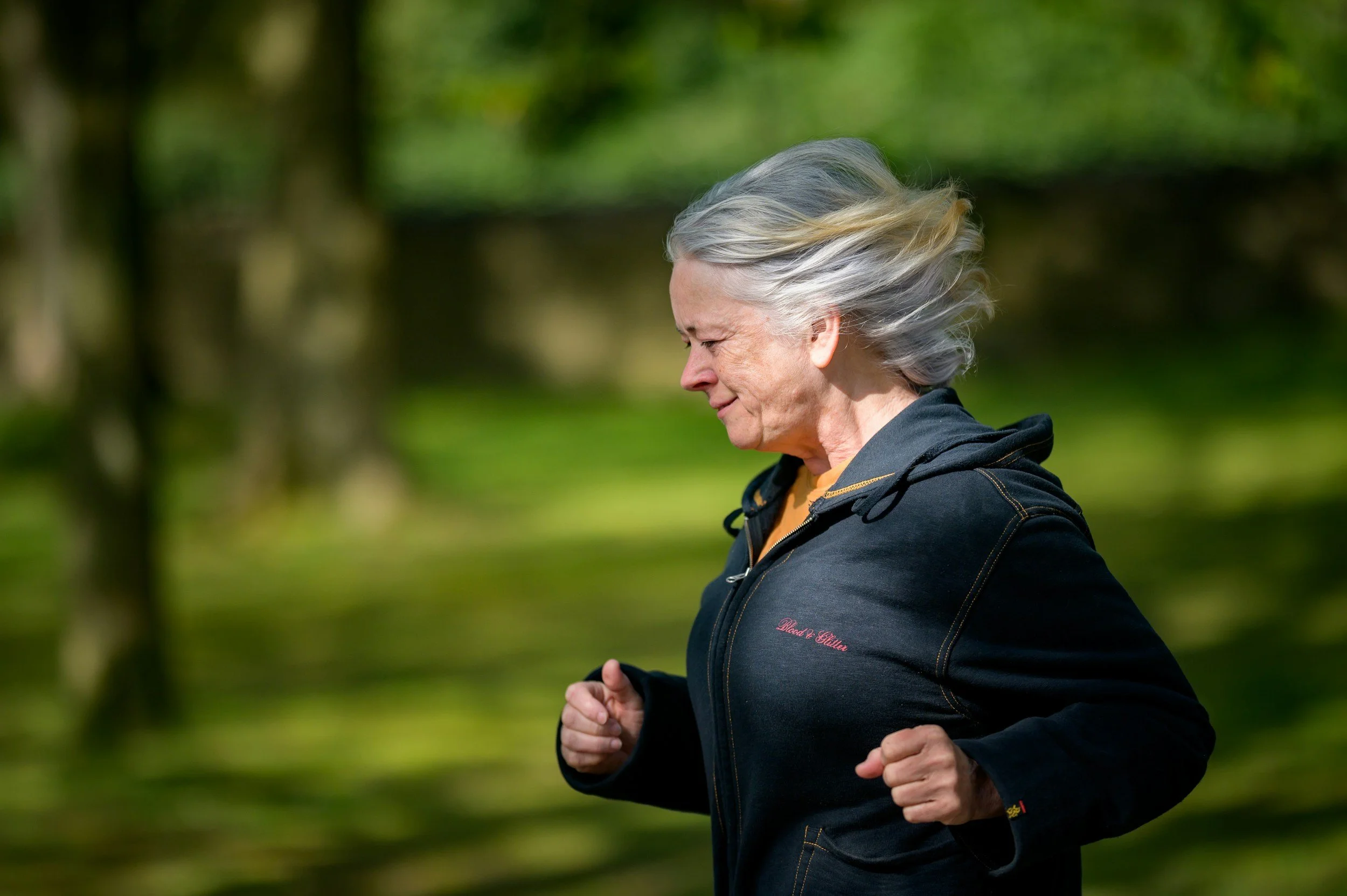 An elderly woman with gray hair running outdoors in a park or wooded area.