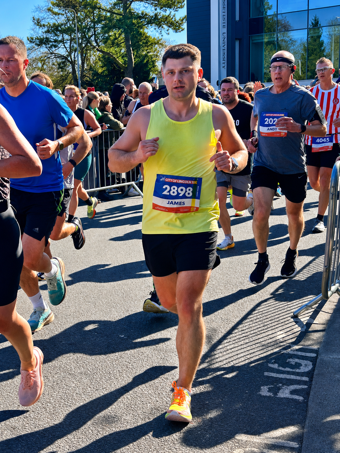 A group of runners participating in a marathon race on a city street, with spectators behind metal barricades and a modern building with reflective windows in the background. The runners are wearing athletic gear and numbered bibs.