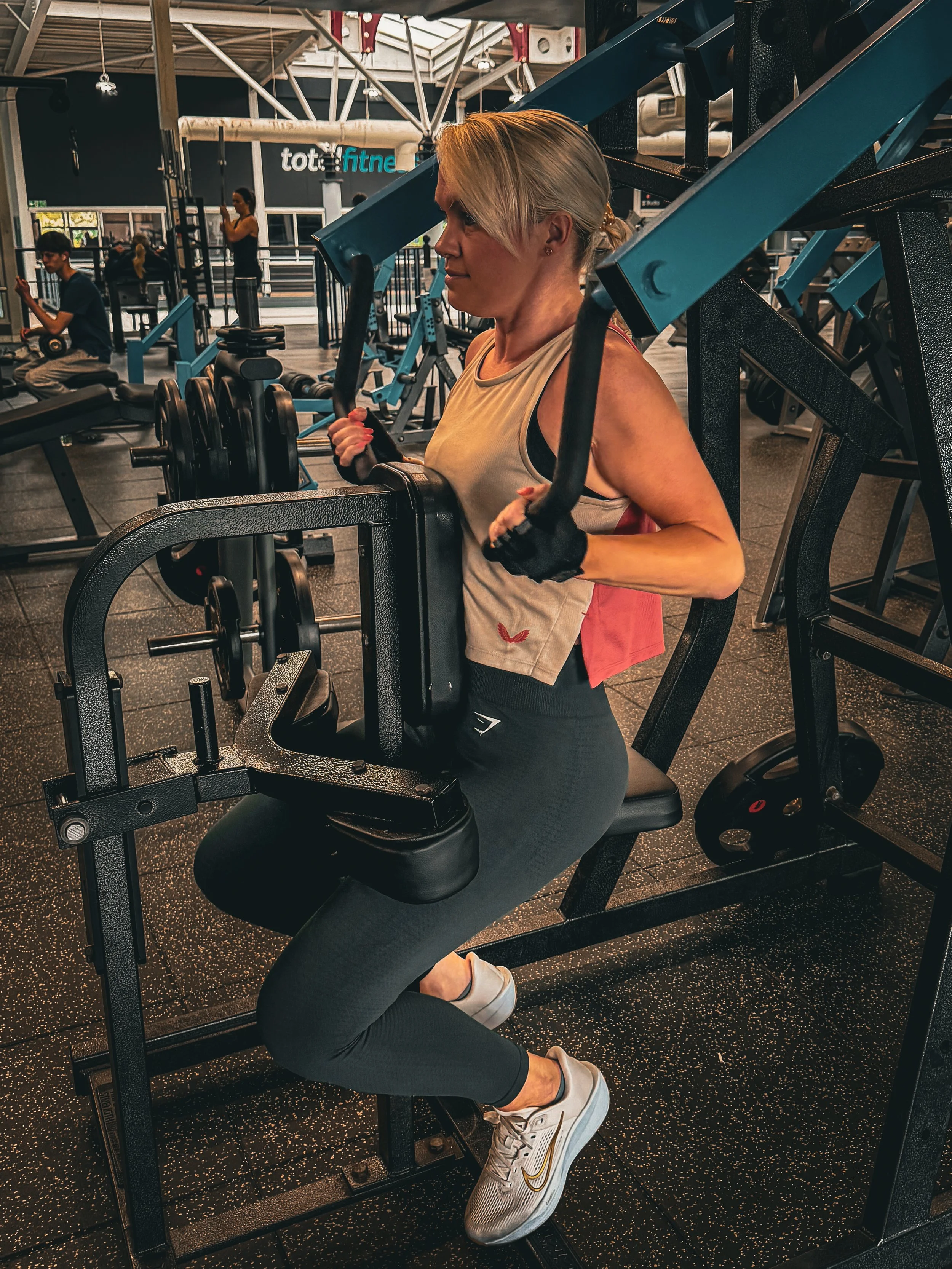 A woman in workout attire performing a leg press exercise at a gym, with other gym-goers visible in the background.