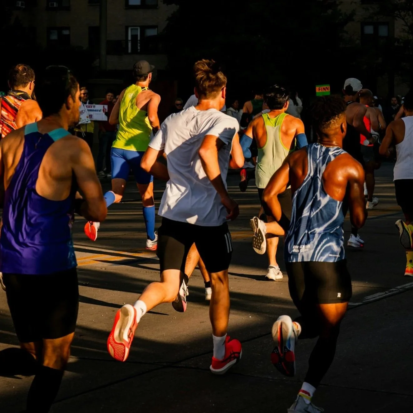 A group of runners participating in a road race during sunset, dressed in athletic gear, running on a city street with buildings in the background.