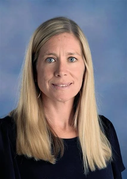 Portrait of a smiling woman with long blonde hair, wearing a black top and hoop earrings, standing against a blue background.