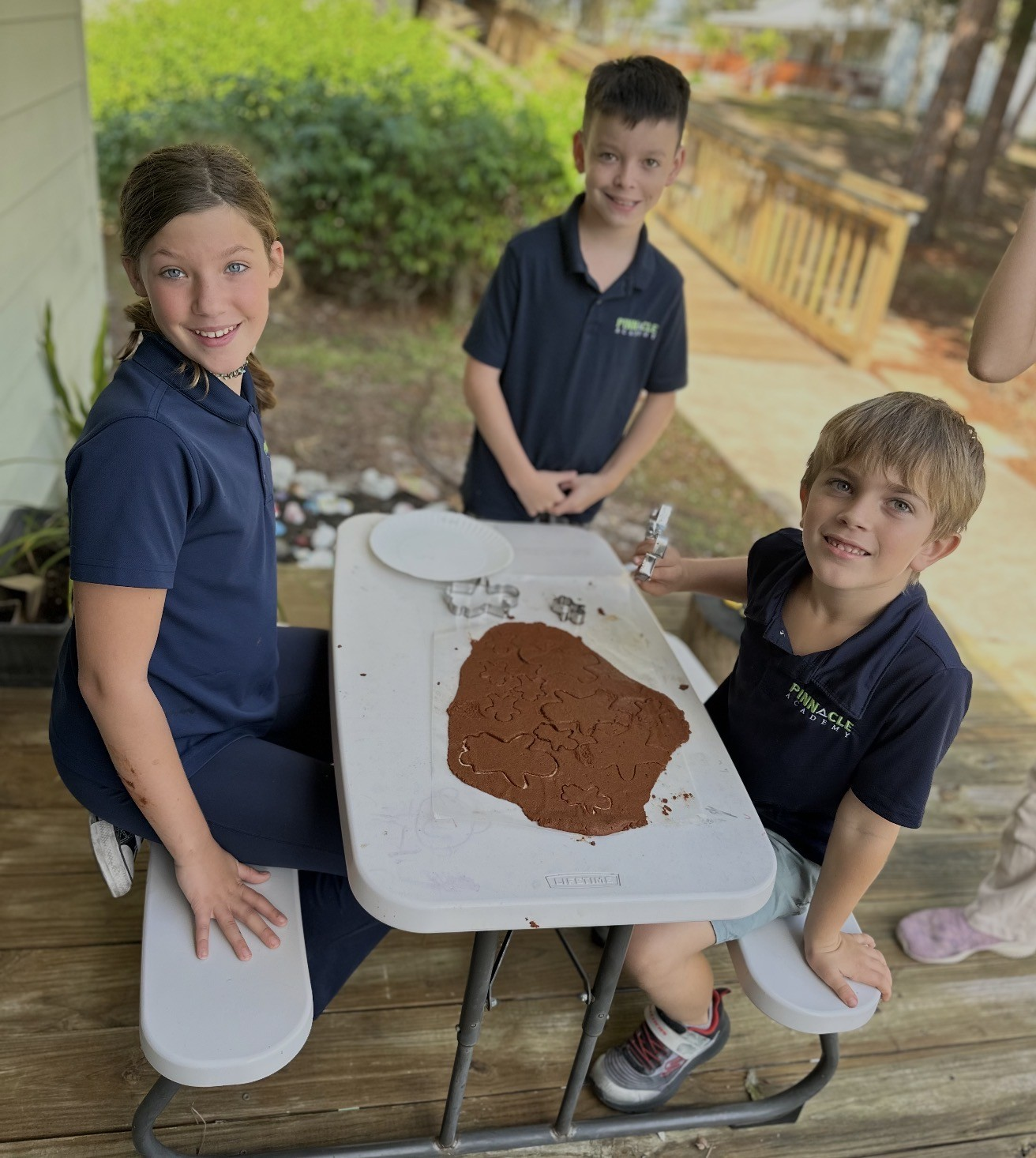 Three children at a table with cookie cutters and rolled-out gingerbread dough, preparing to make cookies outdoors.