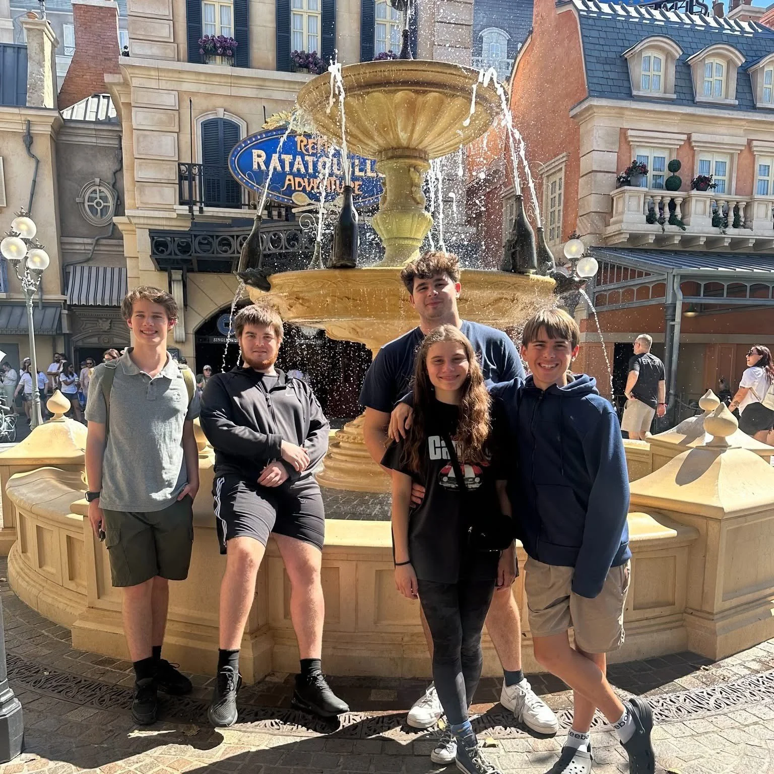 Six teenagers standing in front of a decorative fountain at a theme park, with buildings and other visitors in the background.
