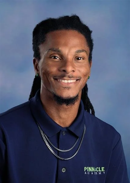 Portrait of a smiling man with dreadlocks wearing a light blue polo shirt with the Pinnacle Academy logo against a blue background.