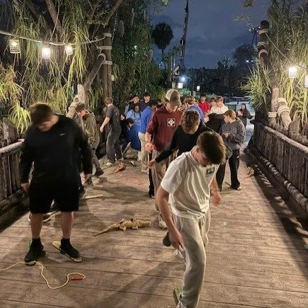 People gathered on a wooden bridge during evening, with some looking down at the ground and others standing, surrounded by tropical plants and illuminated by string lights.