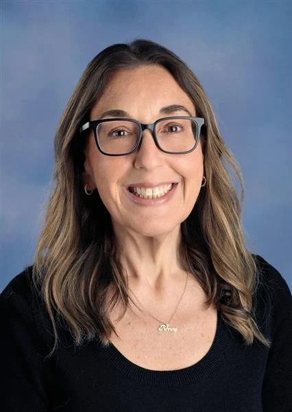 A woman with long brown hair, light skin, wearing a black top and a necklace, smiling at the camera against a blue background.