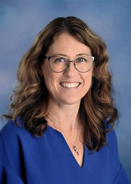 A woman with long curly red hair, wearing glasses and a floral sleeveless top, smiling against a blue background.