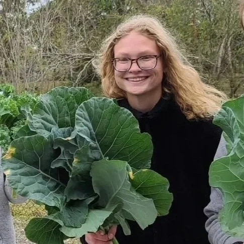 A person with glasses and blonde hair smiling outdoors, holding large green leafy vegetables.