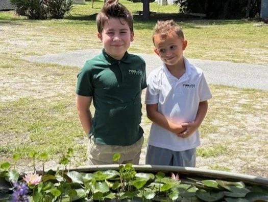 Two young boys standing outdoors near a pond with water lilies, smiling at the camera.