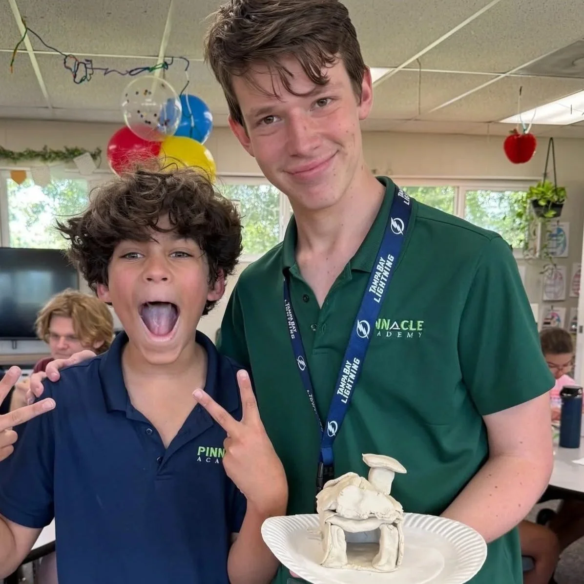 Two boys in a classroom celebrating, with one holding a paper plate with a mushroom-shaped object made of clay or similar material. The boy on the left is making a peace sign and has curly dark hair, wearing a navy polo shirt. The taller boy on the r