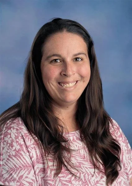 A woman with long dark hair smiling in front of a blue background, wearing a patterned blouse with red and blue floral designs.