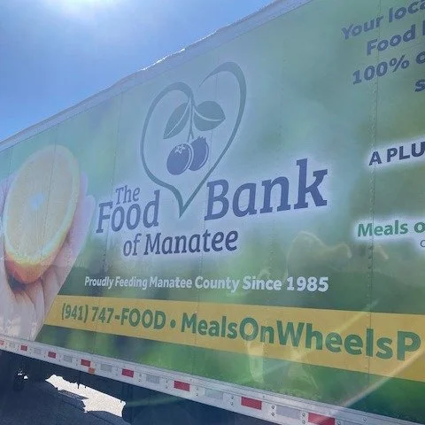 Side of a food truck labeled 'The Food Bank of Manatee,' with a logo featuring a heart and cherry inside a heart. The truck has the tagline 'Proudly Feeding Manatee County Since 1985' and the phone number (941) 747-FOOD, along with the hashtag 'Meals
