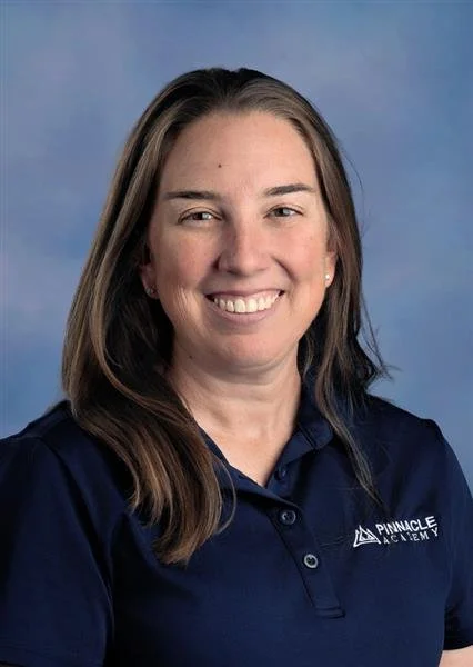 A woman smiling with long brown hair wearing a black polo shirt with a logo on the left chest that reads 'PINNACLE ACADEMY TEACHER' against a blue background.