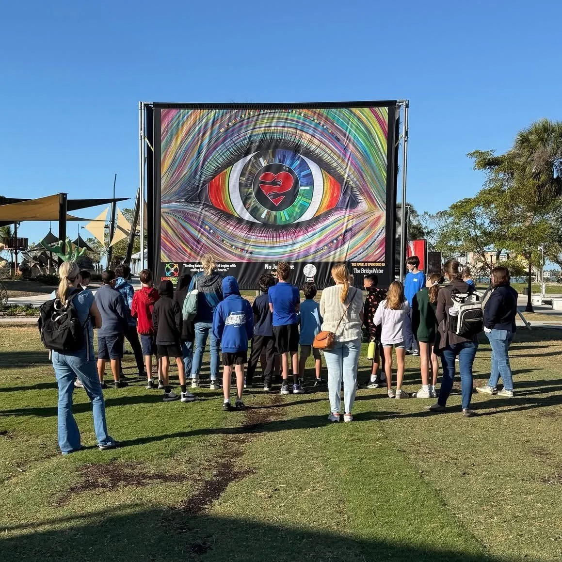 Group of people watching an outdoor movie screen with a colorful eye graphic at a park on a sunny day.