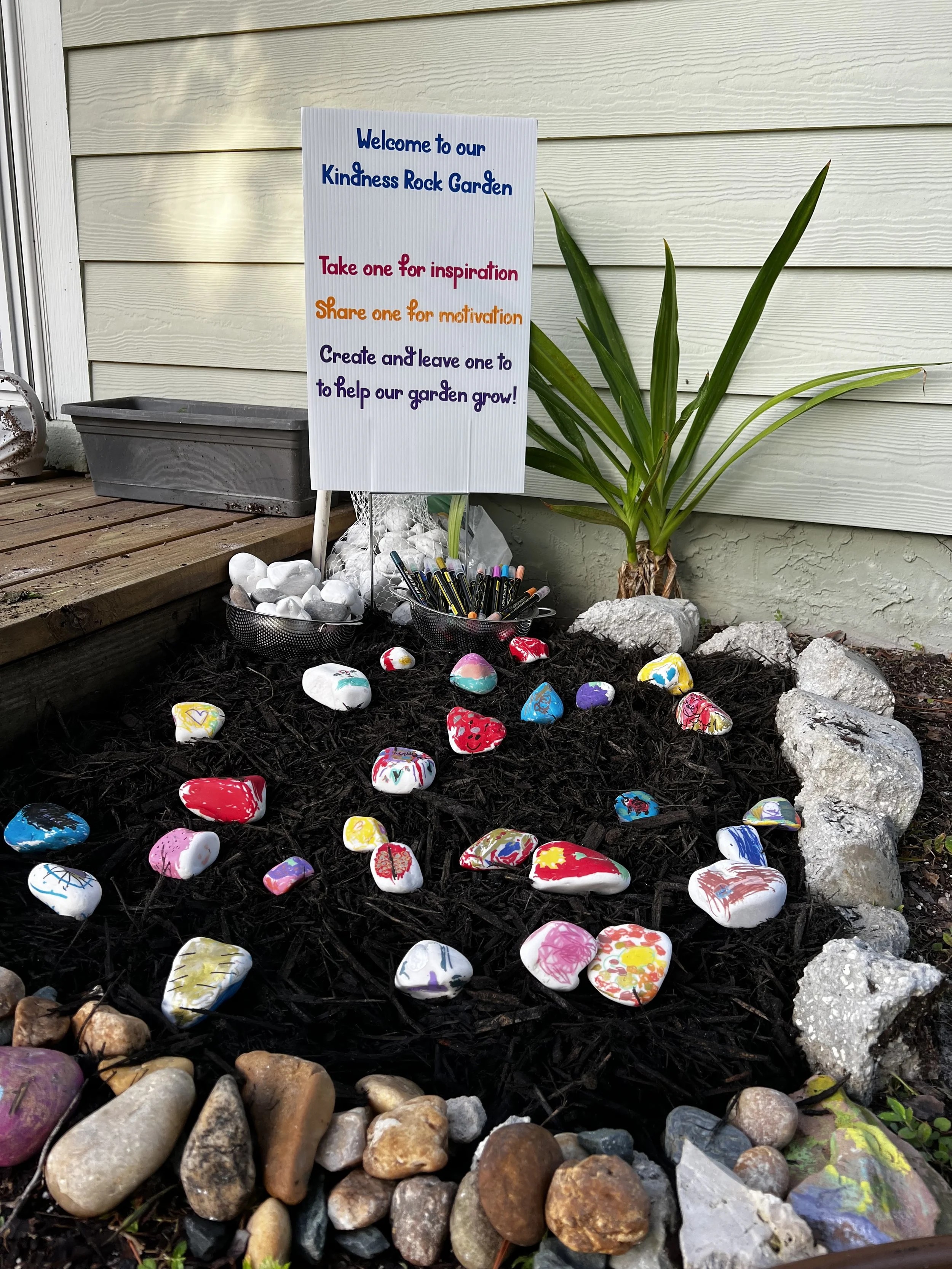 Colorful painted rocks arranged on soil with a sign that reads "Welcome to our Kindness Rock Garden. Take one for inspiration. Share one for motivation. Create and leave one to help our garden grow!" and a plant with long green leaves next to the sign.