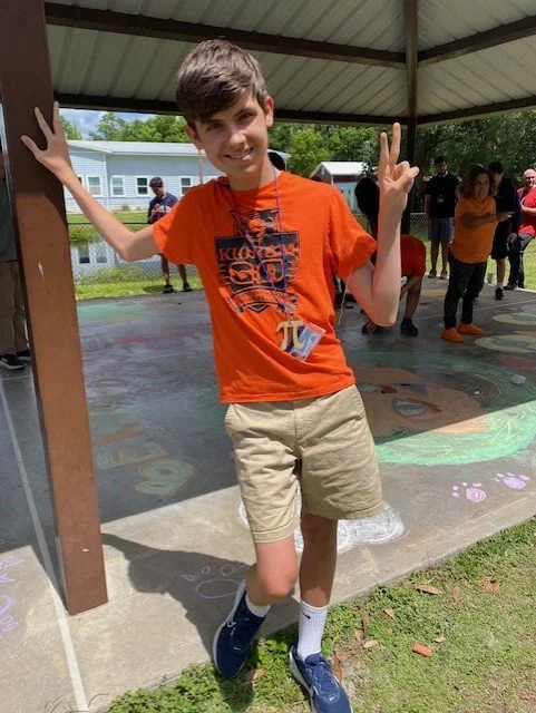 Smiling boy in orange shirt and beige shorts making peace signs at an outdoor gathering under a pavilion.
