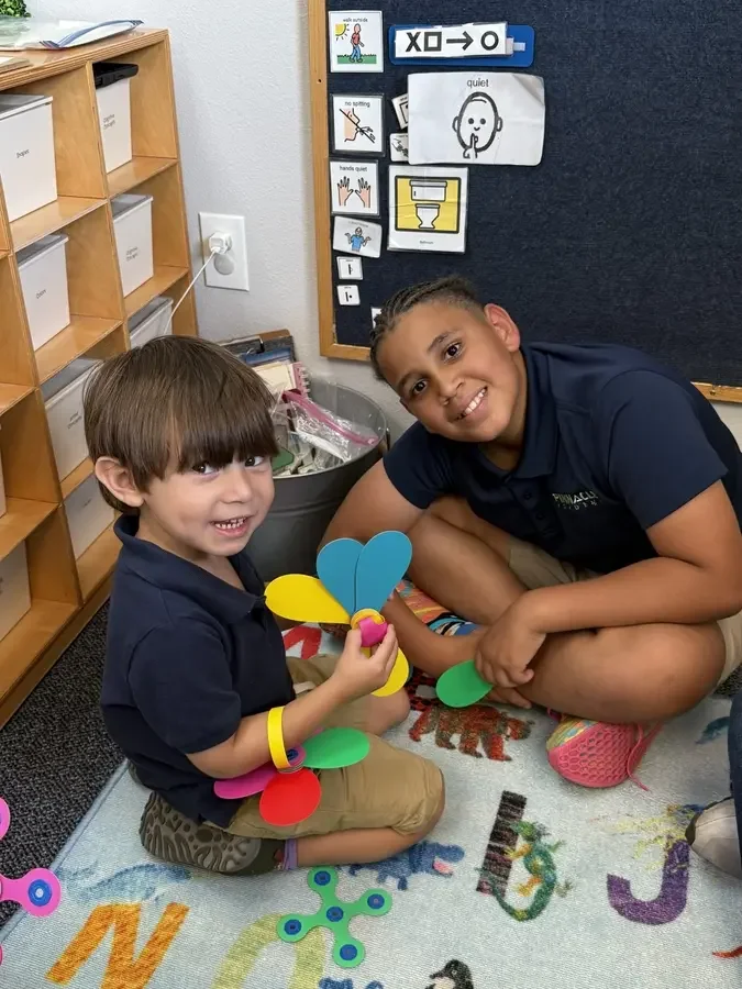 Two children sitting on a colorful alphabet rug at school, smiling, one holding colorful butterfly craft materials, and the other leaning next to them.