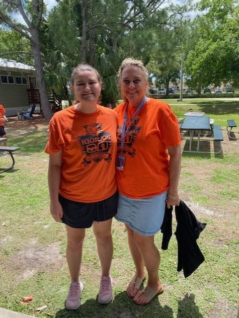 Two women standing outdoors on grass, both wearing orange T-shirts with a logo, smiling, with trees and picnic tables in the background.