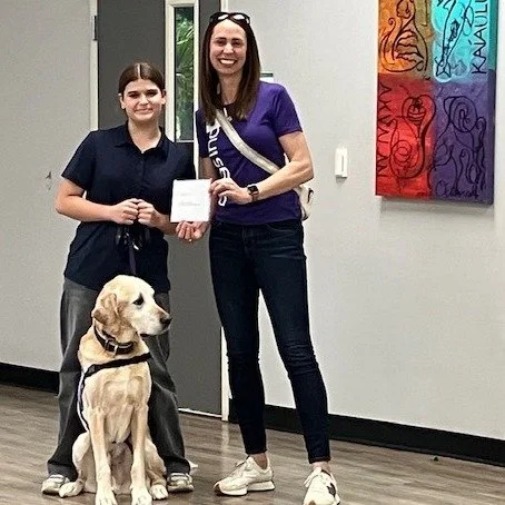 Two women and a guide dog in an indoor setting, with one woman handing a paper to the other, and a large Labrador sitting beside them.