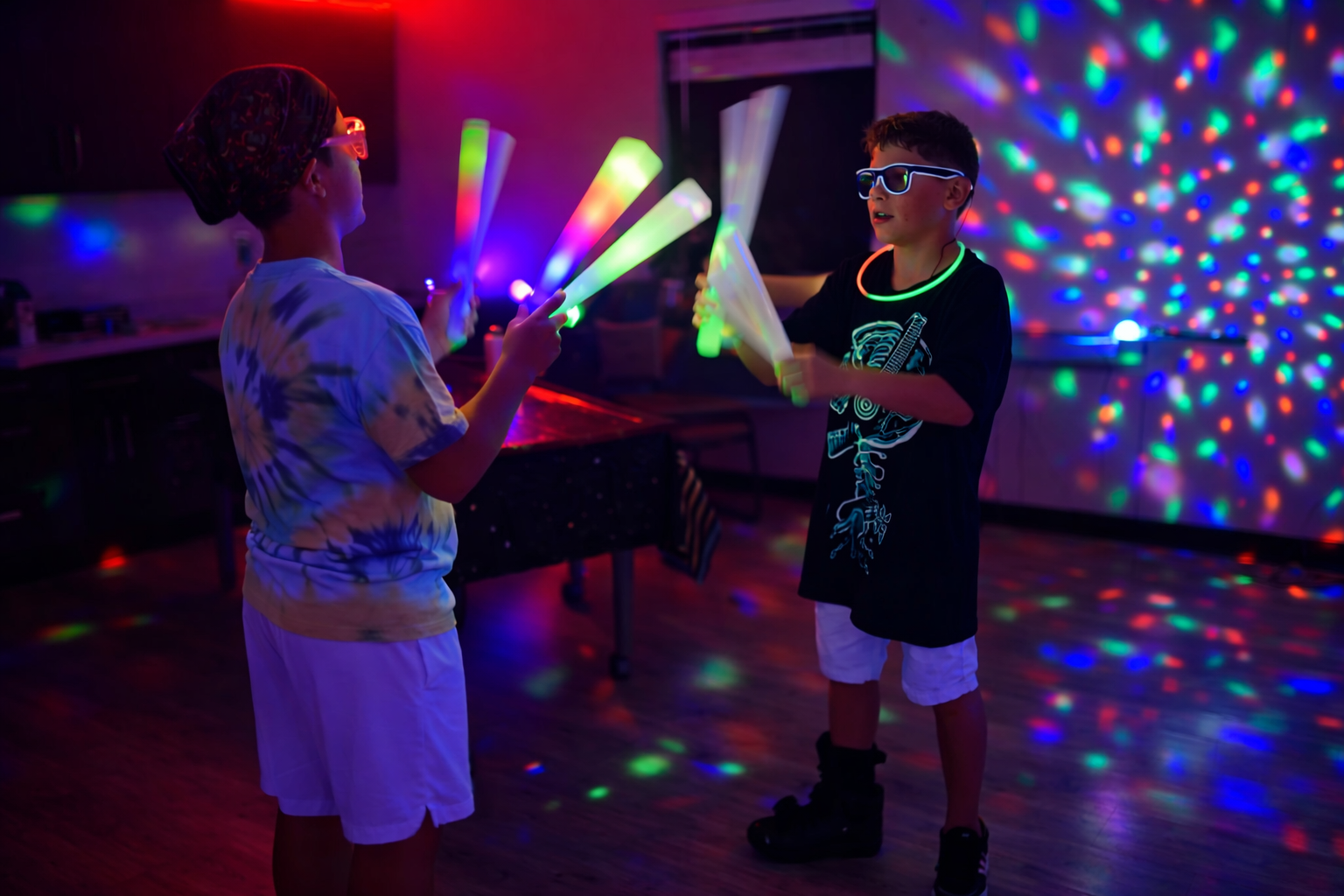 Two children at a glow party are wearing glasses and glow necklaces, holding glow sticks, with colorful light projections on the wall in the background.