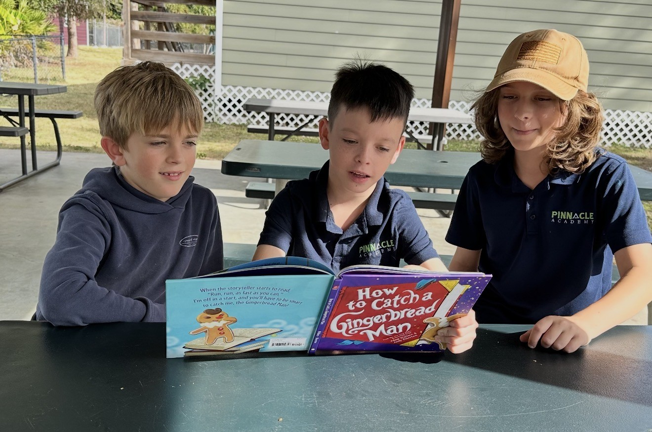 Three boys in navy shirts reading a book titled 'How to Catch a Gingerbread Man' at a table outdoors.