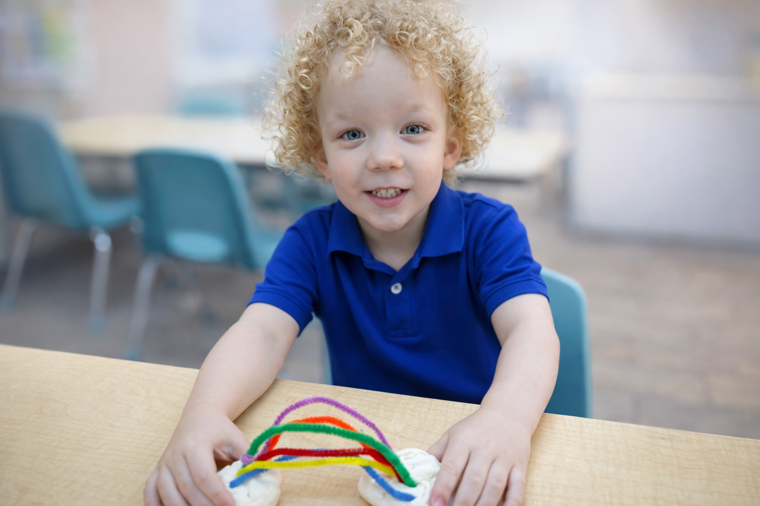 A young boy with curly blond hair and blue eyes, wearing a blue polo shirt, is sitting at a table with colorful pipe cleaners on it. The background features a blurred classroom setting with blue chairs and tables.