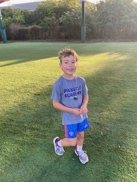 A young boy smiling and running on a grassy field during daytime, wearing a Pinnacle Academy t-shirt, blue shorts, sneakers, and a medal around his neck.