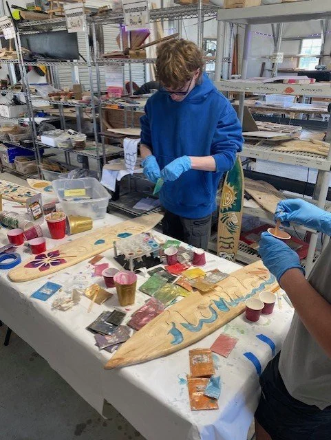 Two people working on art projects at a workspace with craft supplies, including paint, brushes, and surfboard-shaped wooden pieces.