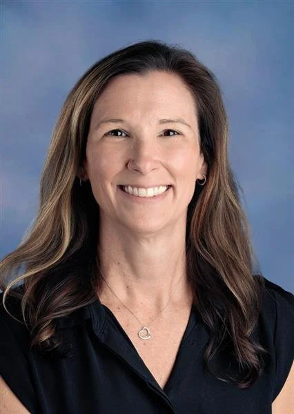 A woman with long brown hair wearing a black sleeveless top and a silver necklace with a heart-shaped pendant, smiling against a blue background.