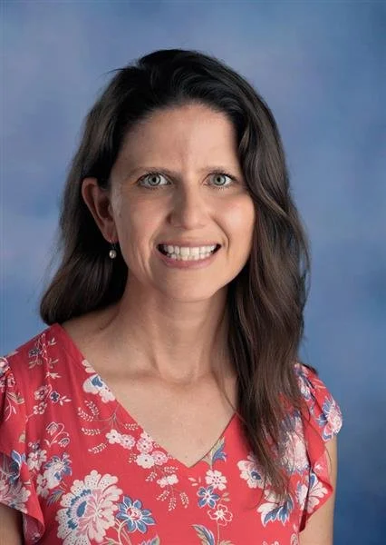 A woman with shoulder-length brown hair smiling against a blue background, wearing a black sleeveless dress with a white floral pattern and pearl earrings.