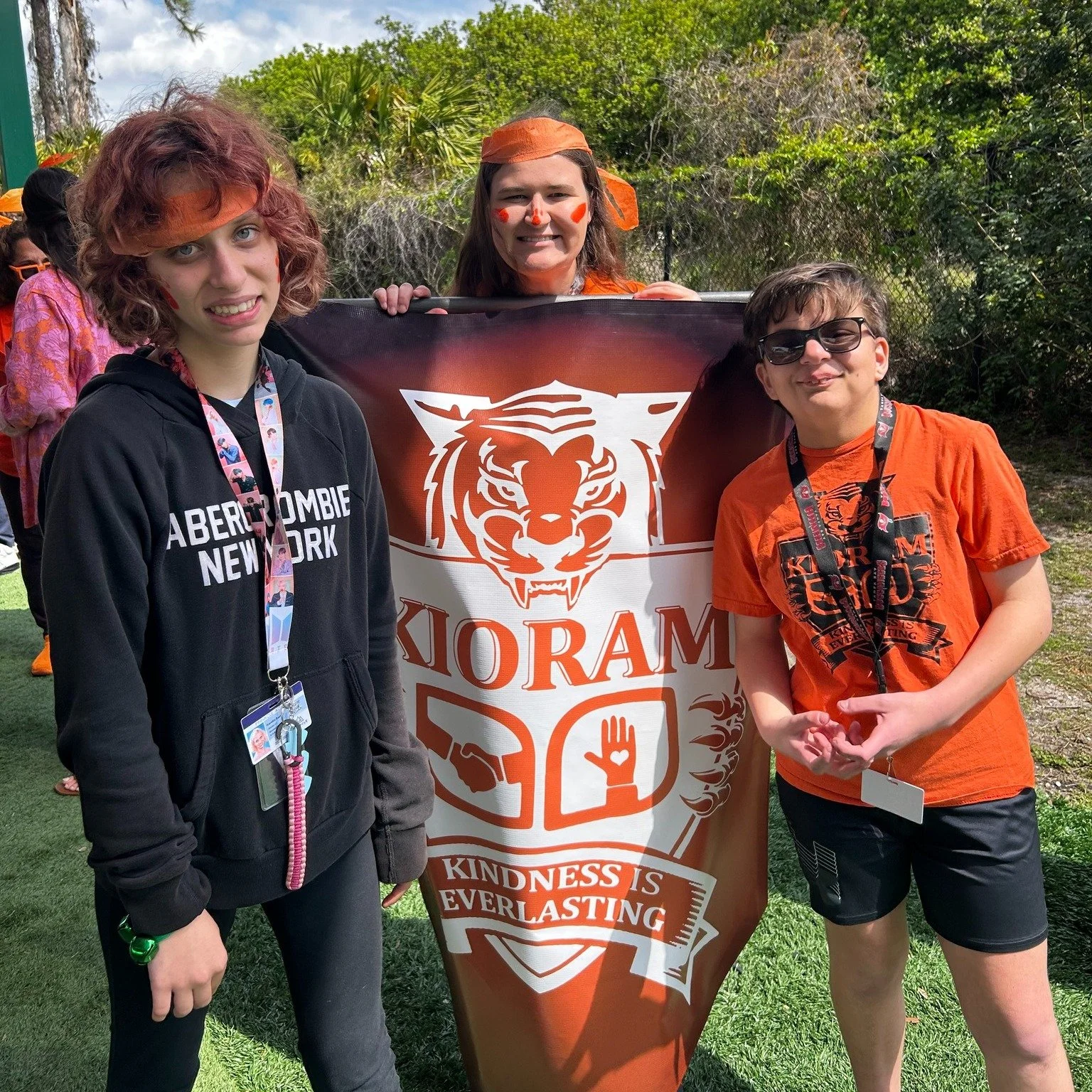 Four people, including three children, standing outdoors holding a large banner with a tiger logo and the words 'Kindness is Everlasting' on it. They are dressed casually, and the background features trees and a sunny sky.