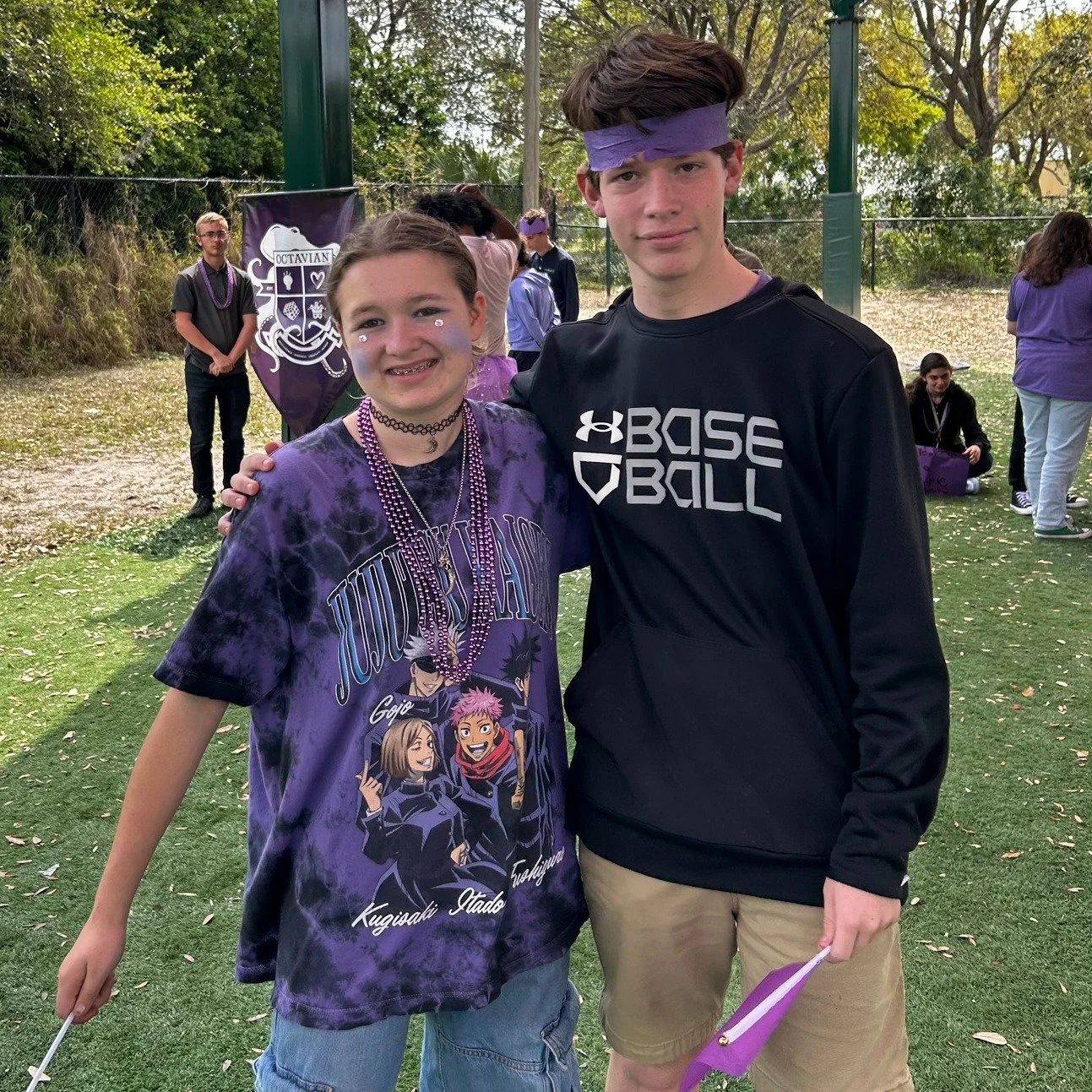 A teenage girl and boy at an outdoor event, smiling and standing close together, with the girl wearing a purple tie-dye JuJutsu T-shirt and the boy wearing a black Under Armour football sweatshirt.