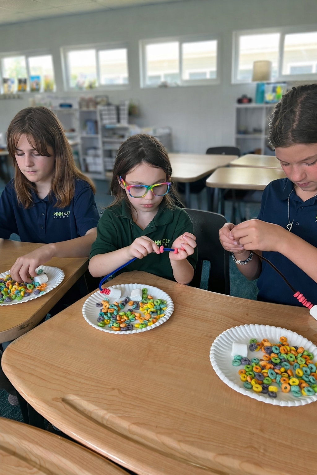 Three children sitting at a table, working with colorful cereal and marshmallows on paper plates, in a classroom setting.