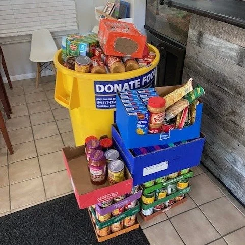 Multiple cans and boxes of food stacked next to a donation bin labeled 'Donate Food' in a room with tiled floor and a small stool nearby.
