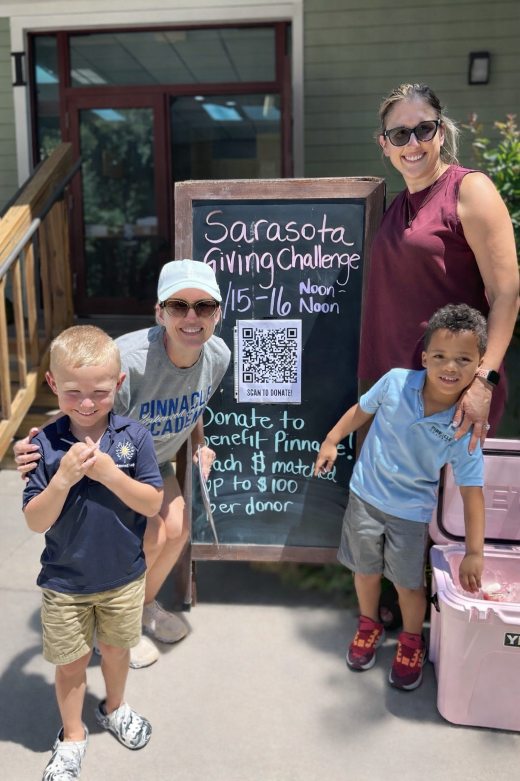 A group of four people, including two young boys and two women, standing outside in front of a chalkboard sign that reads 'Sarasota Giving Challenge, 1/15-1/16, Noon-12 Noon,' during a charity event. One woman is crouching and wearing a white cap and sunglasses, while the other woman is standing and wearing sunglasses and a sleeveless maroon top. The boys are smiling, with one amassing in a blue polo and the other in a light blue polo. There is a QR code on the sign for donations.