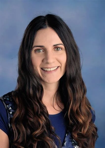 A woman with long brown hair wearing a black sleeveless top, smiling against a blue background.