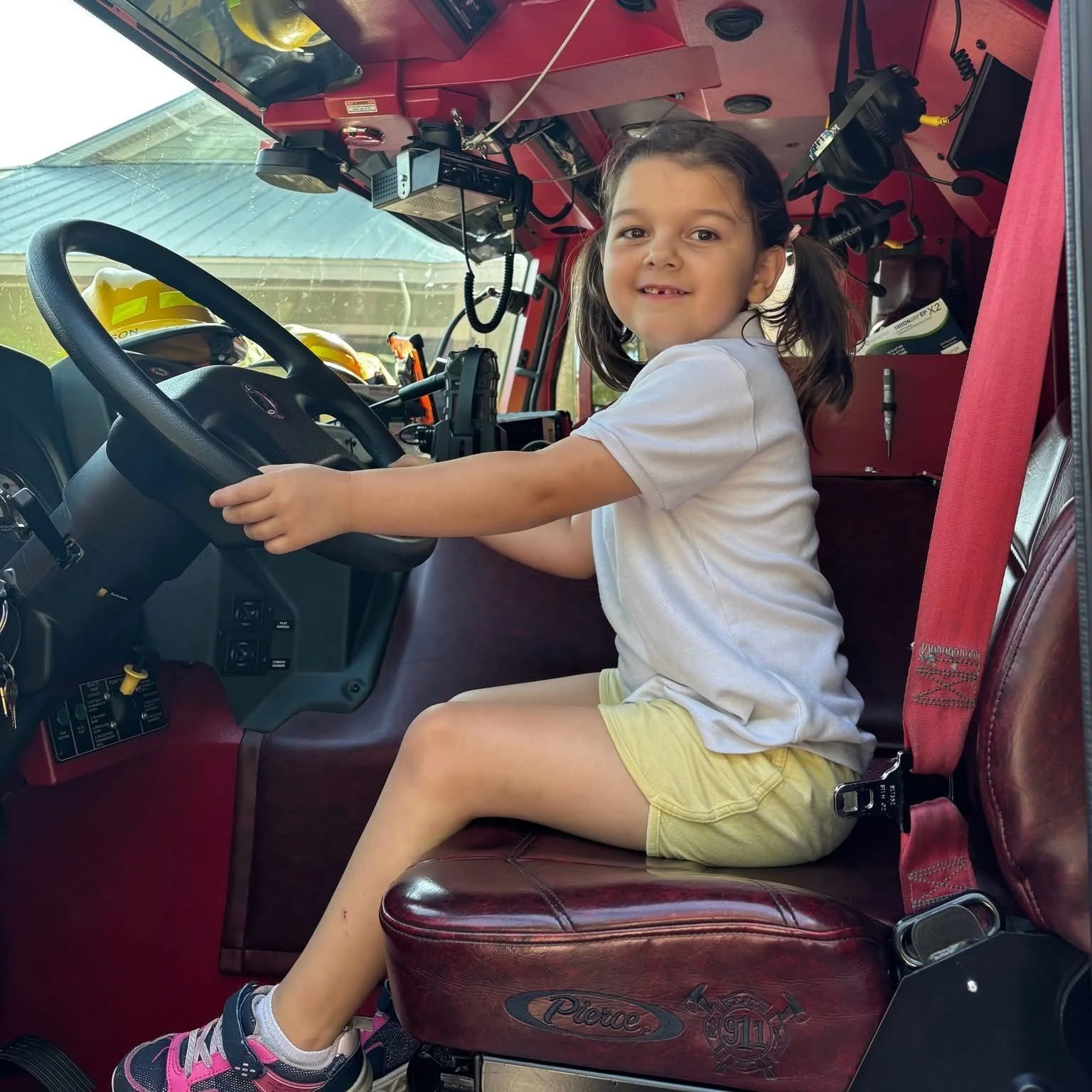 A young girl sitting in the driver's seat of a fire truck, smiling at the camera, with firefighter helmets visible on the dashboard.