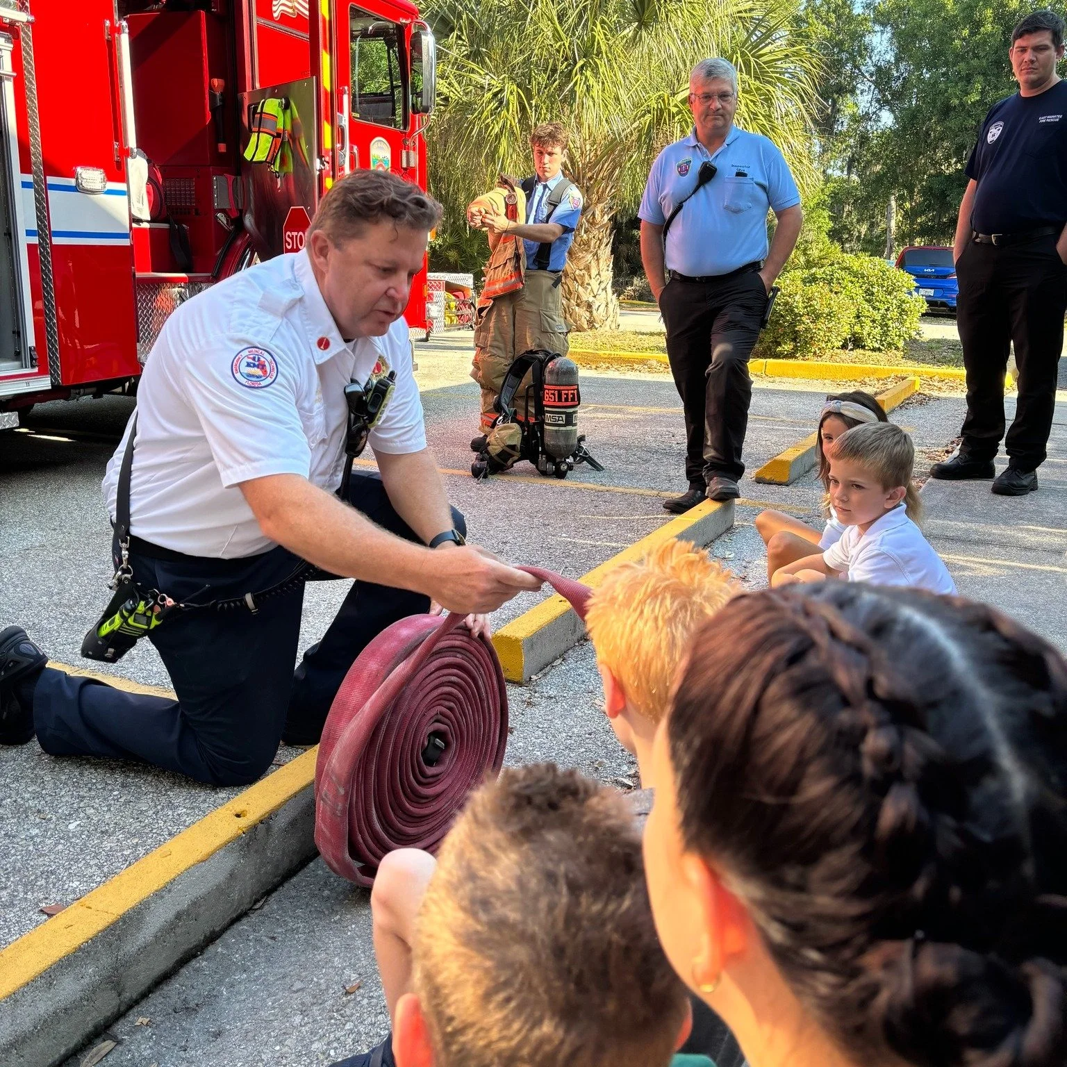Firefighter kneeling on the ground and holding a fire hose, demonstrating to a group of children and adults in a parking lot, with a fire truck and palm trees in the background.