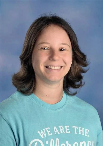 Young woman with shoulder-length light brown hair and hoop earrings smiling, wearing a sleeveless beige top with circular patterns, standing in front of a blue background.