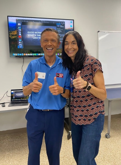 Two smiling people, a man in a blue shirt and a woman in a patterned blouse, standing in front of a large screen and giving thumbs up.