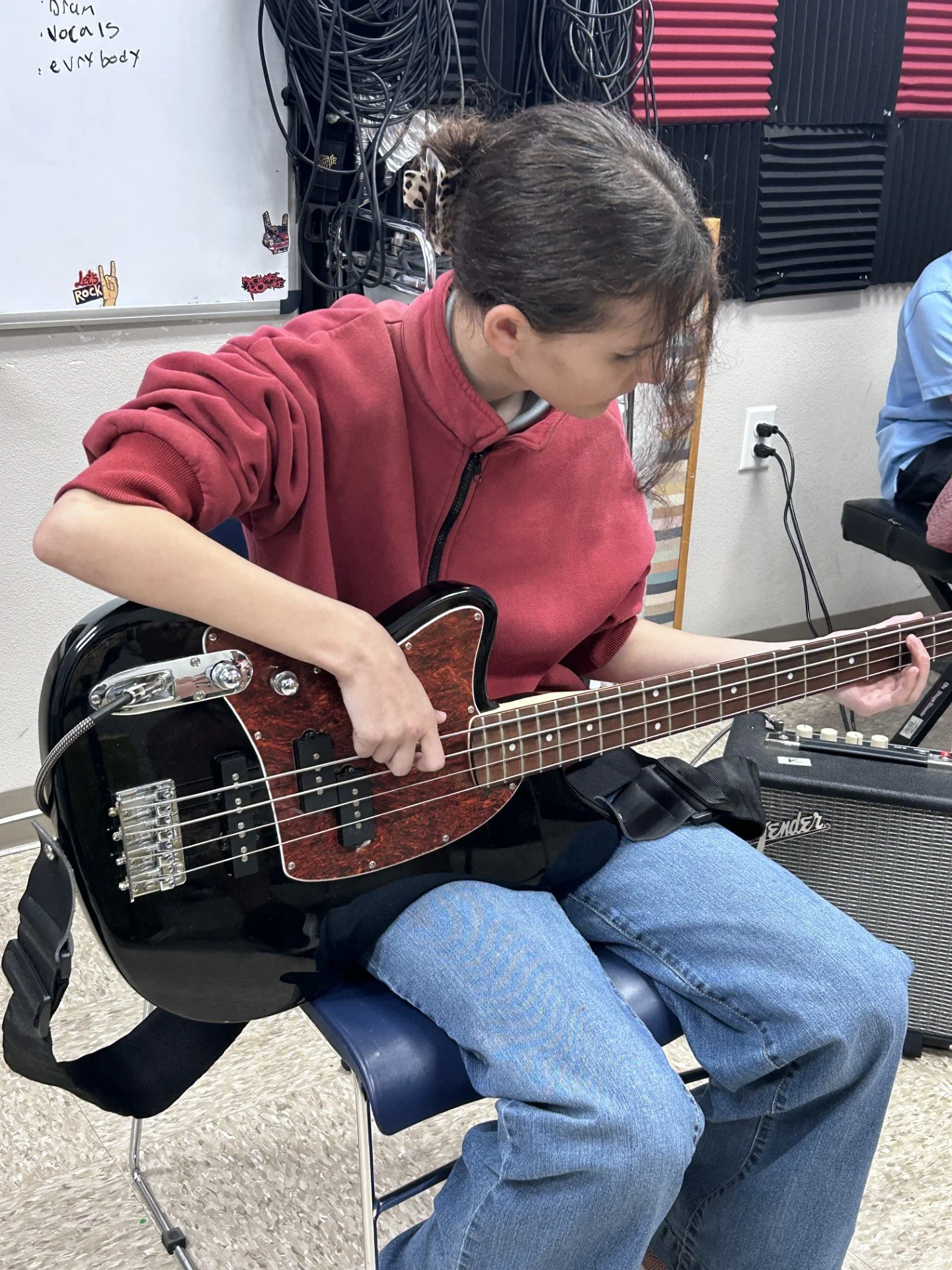 Person sitting on a chair playing a bass guitar, wearing a red hoodie and blue jeans, in a music room with soundproofing foam and a whiteboard.