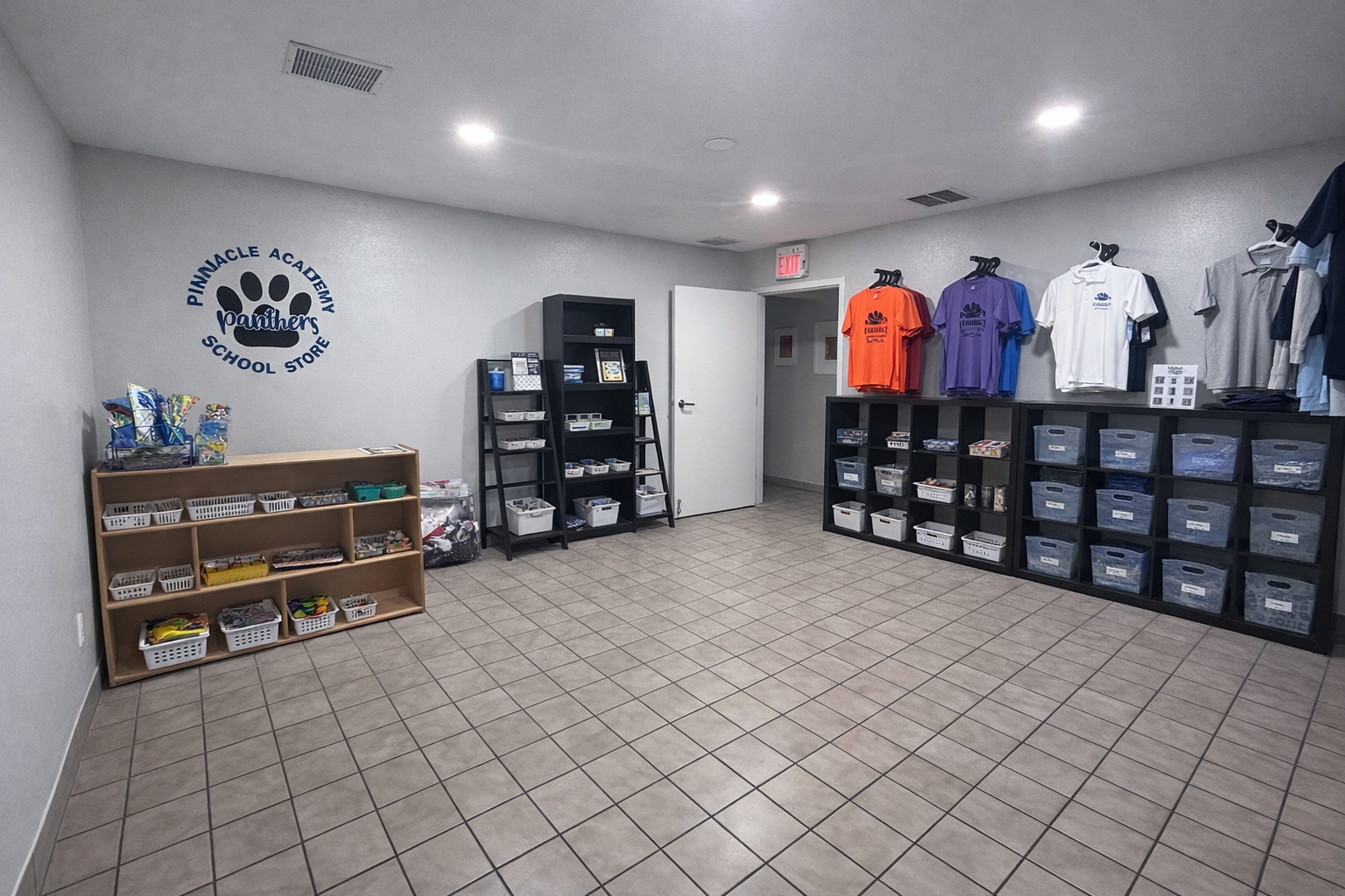 An indoor school store with a gray wall featuring Pinnacle Academy Panthers logo, shelves with school supplies and clothing, and a tiled floor.